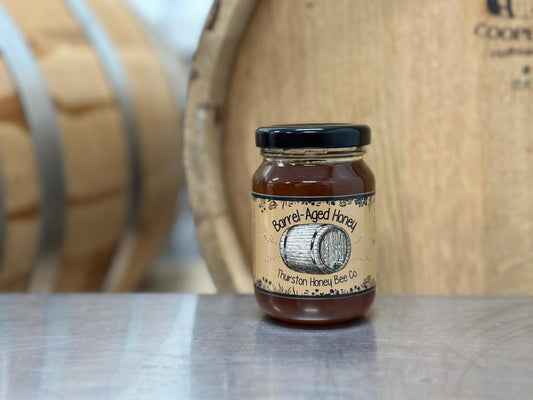 Jar of barrel-aged honey on a metal surface with wooden barrels in the background